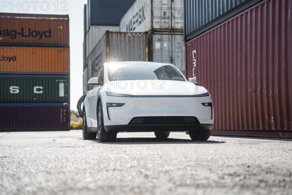 White car in a harbour environment next to stacked freight containers, Tesla Model Y Juniper,