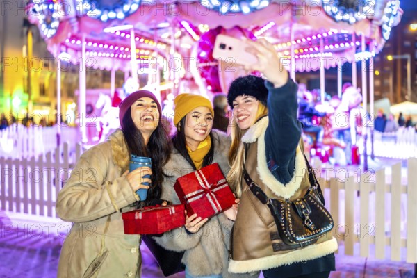 Three cheerful young women taking a selfie at a christmas market, holding gifts and warm drinks,