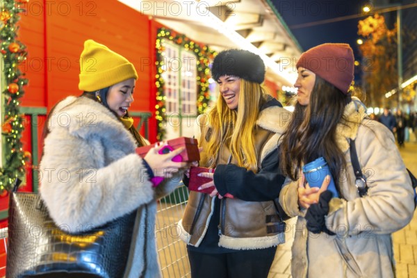 Three cheerful young women exchanging christmas presents at a decorated outdoor night market,