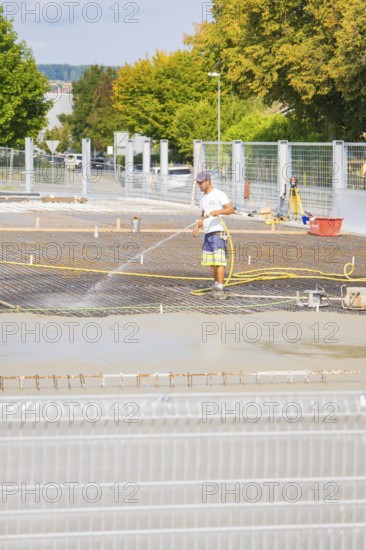 Construction worker watering concrete on a building site, surrounded by safety fences, car park