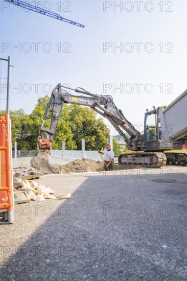 A large excavator and a construction worker on a building site with gravel and a lorry. Blue sky