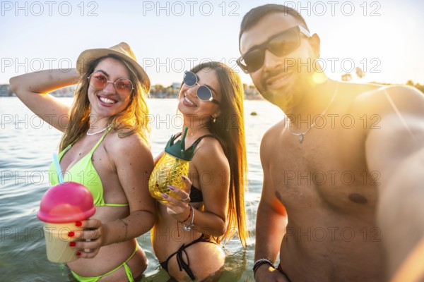 Three cheerful friends are taking a selfie in the sea, holding refreshing cocktails, enjoying a