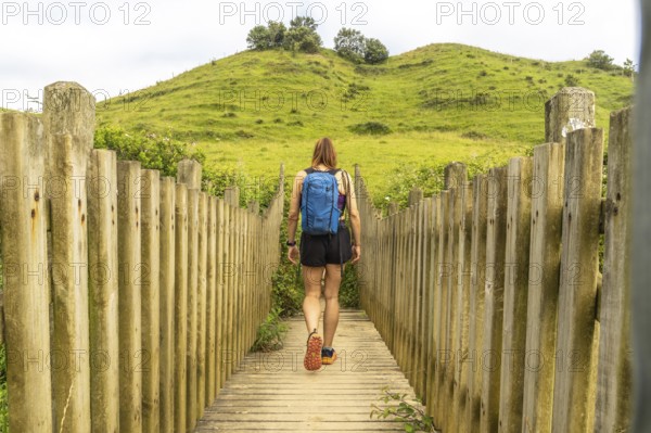 Female hiker with backpack walking on wooden footbridge leading towards a green hill in zumaia,