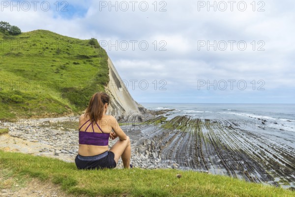 Woman sitting on green grass overlooking the impressive flysch cliffs and coastline of sakoneta