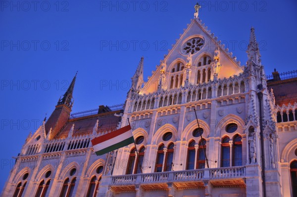 Ornate facade of hungarian parliament building illuminated at twilight with hungarian flag waving,