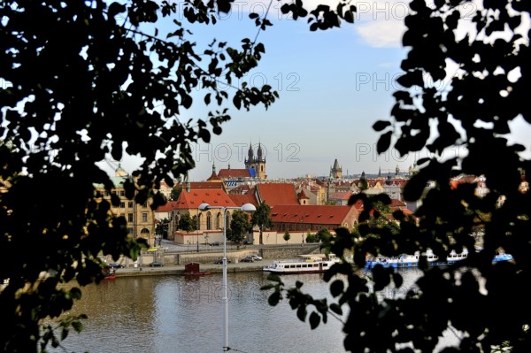 Prague cityscape with vltava river, church of our lady before tyn and tiled roofs framed by leaves