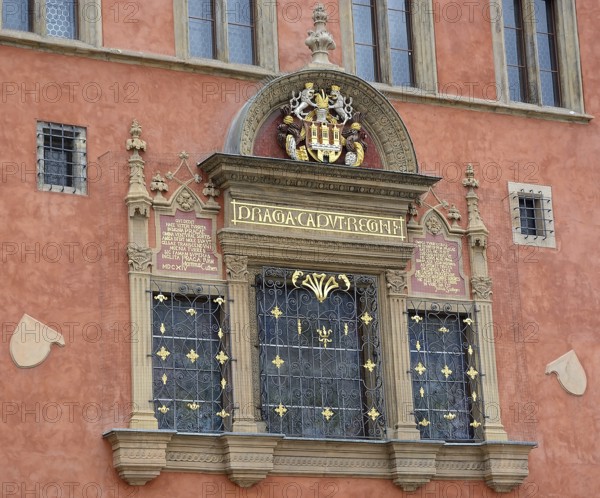 Architectural detail showcasing an ornate window with a coat of arms, latin inscription, and