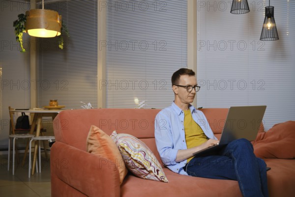 A man sits comfortably on a sofa and chats online on his laptop during evening hours. He