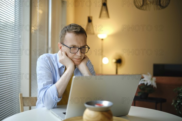 A middle-aged man with glasses watching series on his laptop while seated at a round table in a