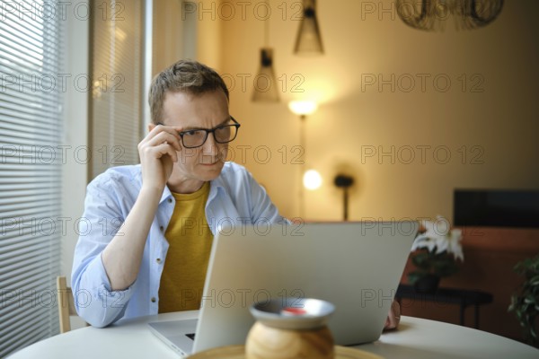 A focused man with glasses is working on his laptop with financial documents at home in the evening
