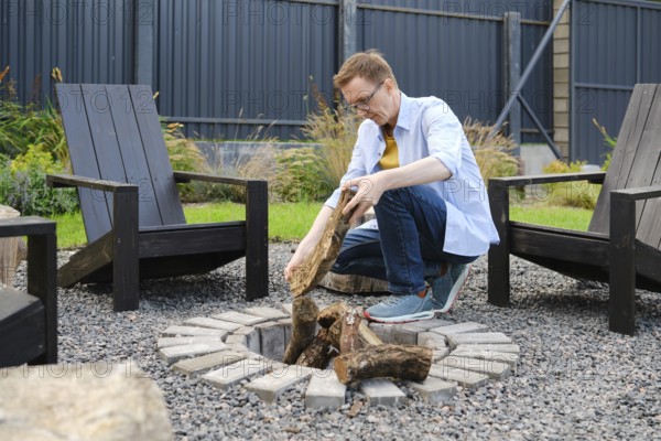 A middle-aged man is kneeling beside a circular stone fire pit, carefully placing logs into the
