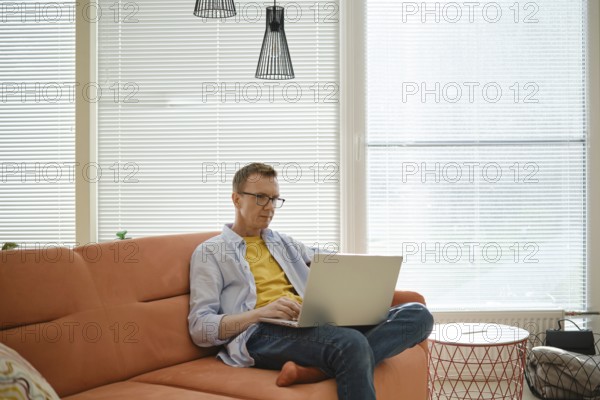 A man is seated on an orange couch, focused on his work on a laptop. Sunlight streams through the