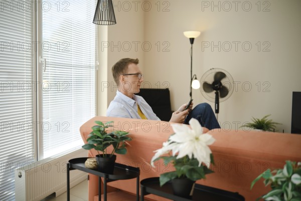 A man sits comfortably on a couch in a modern living room with large windows and natural light. He