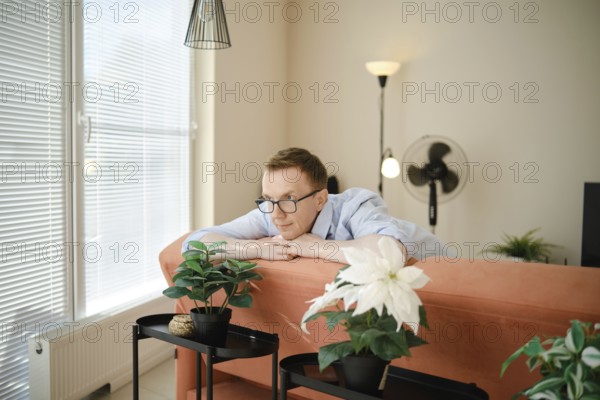A middle-aged man leans on a colourful couch, gazing thoughtfully at potted indoor plants nearby.