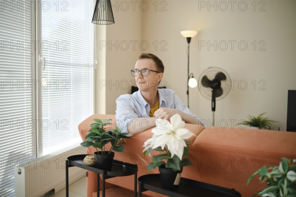 A middle-aged man sits comfortably on a stylish couch, looking contemplatively out of large windows