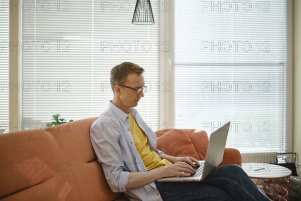 A retired man sits comfortably on a couch and writes his memoirs on laptop in a well-lit living