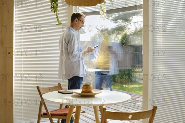 A man stands inside a stylish room, using his smartphone while looking out through a large glass