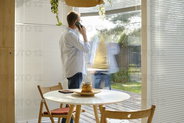 A middle-aged man stands near a large window, engaged in a phone conversation. Sunlight filters