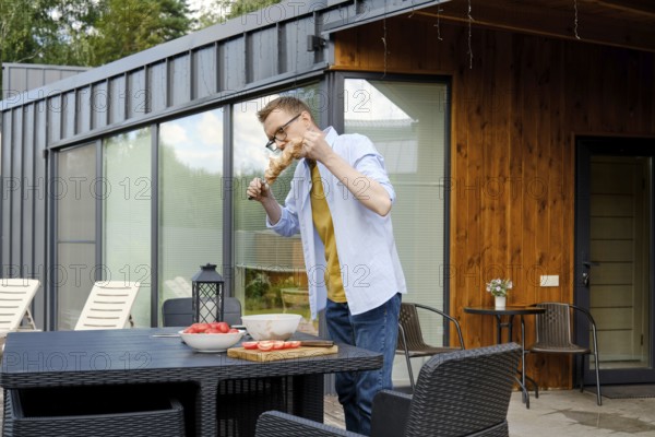 A man is enjoying a summer barbecue at a stylish wooden house. He is focused on grilling meat,