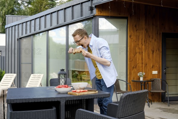 A middle-aged man stands at a patio table of tiny house and playfully bites marinated pork meat on