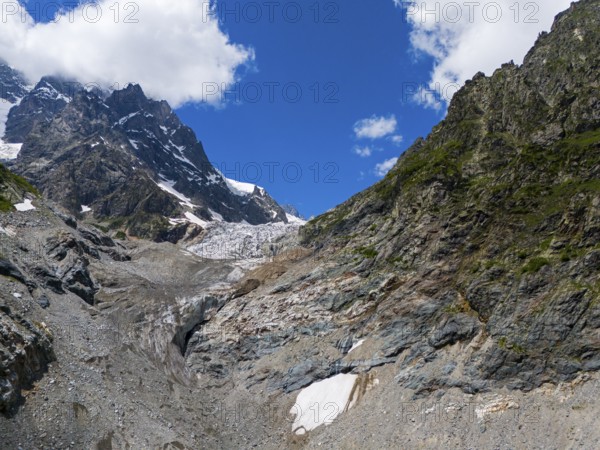 Rocky landscape with a glacier under a blue sky, flanked by mountains. Clouds drift over the alpine
