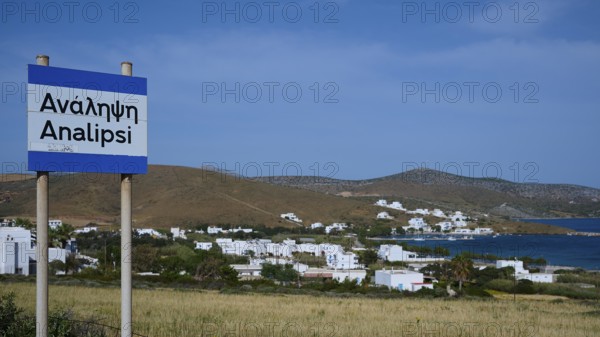 A town sign in front of a coastal settlement with whitewashed houses and blue sky, Analipsi,