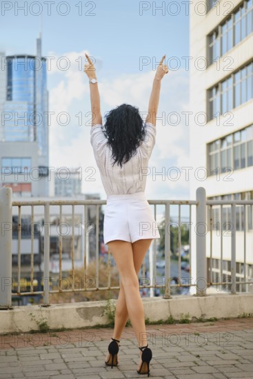 A woman stands outdoors in a vibrant urban setting with arms lifted to the sky. Dressed in a