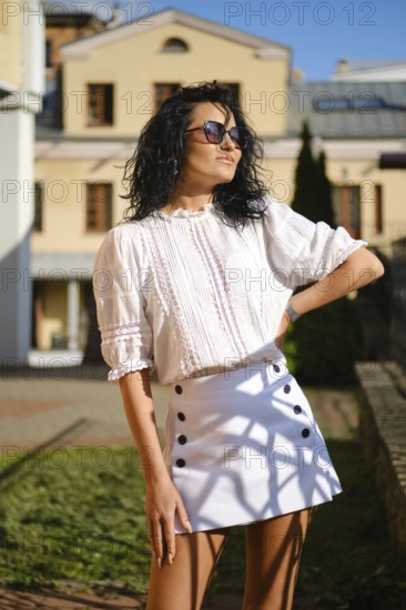 Bright sunlight highlights a woman posing confidently in a chic white blouse and skirt. She stands