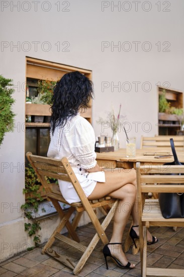A woman with curly hair sits at a wooden table in a rustic outdoor cafe. She wears a white blouse