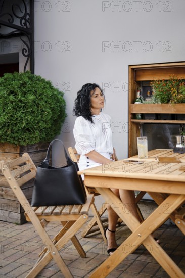 A woman with curly hair relaxes at a wooden table in an outdoor cafe, dressed in a fashionable