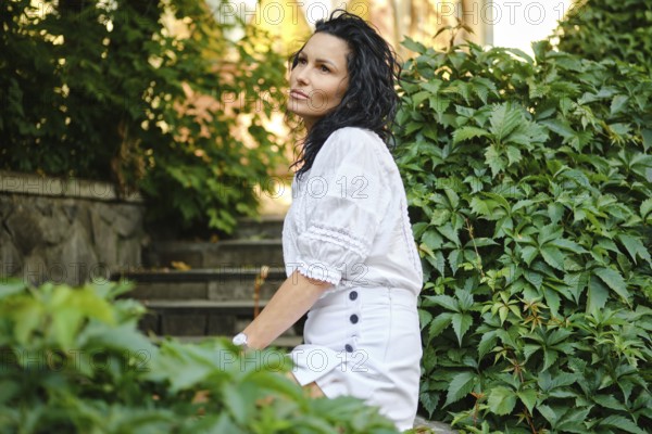 A woman with curly hair is seated gracefully on stone steps surrounded by vibrant green plants. She