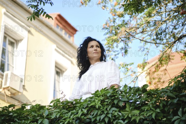 A woman with dark, wavy hair gazes thoughtfully while surrounded by lush green leaves in a charming