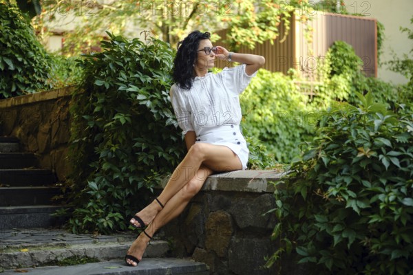A woman with curly hair is seated on a stone wall, wearing a stylish white outfit and sunglasses.