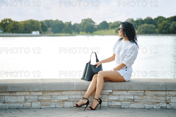 A woman wearing a chic white outfit and black heels relaxes on a stone ledge near calm water. The