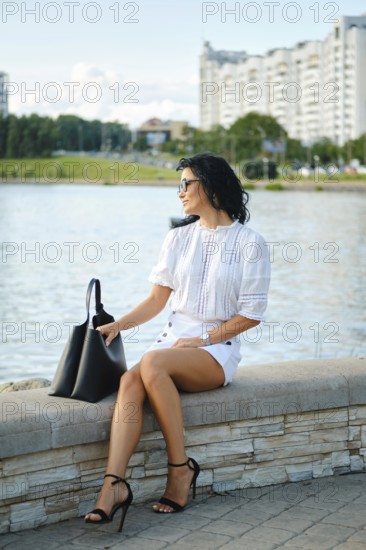 A fashionable woman sits by the water, dressed in a white top and shorts while wearing stylish