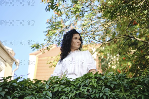 A woman with dark, curly hair stands amidst lush green leaves, looking thoughtfully into the