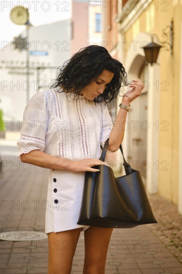 A woman with curly hair stands in a charming alleyway, wearing a white blouse and a stylish skirt.