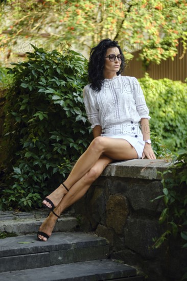 A woman sits elegantly on a stone ledge in a garden, dressed in a stylish white top and mini skirt.