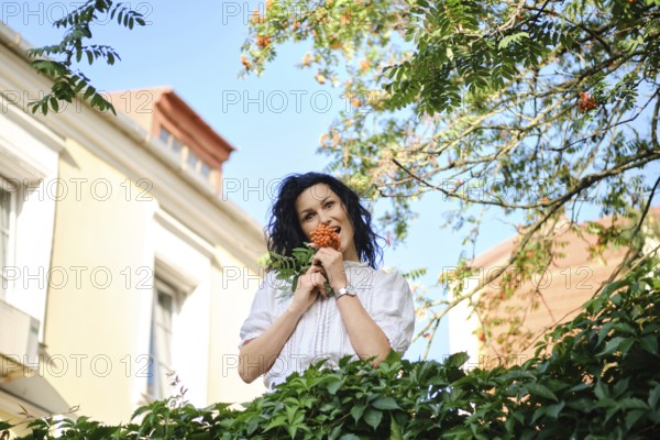 A playful woman stands surrounded by greenery holding a cluster of bright berries. The sun shines