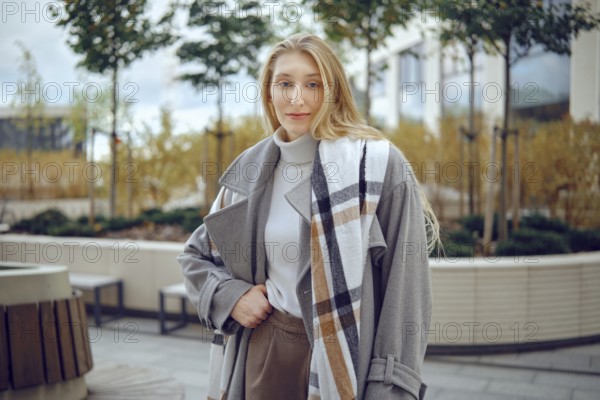 A young woman stands outdoors, showing off her fashionable gray coat featuring a plaid pattern. She