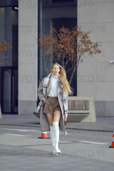 A confident woman strolls along a city street, wearing a trendy autumn outfit that includes boots