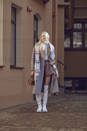 A fashionable young woman in wool coat, knee-high boots and scarf posing in a sunny urban alley.