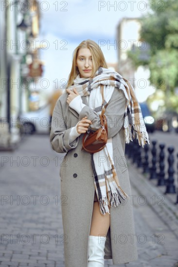 A young woman with blonde hair stands on a cobblestone street, wearing a fashionable coat and a