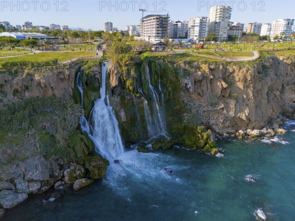 A waterfall cascades from a cliff into the sea, surrounded by urban landscape and green parks,