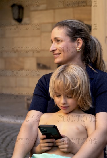 Mother, woman, has boy, 6 years, blonde, in arms, playing with smartphone, mobile phone, upper body