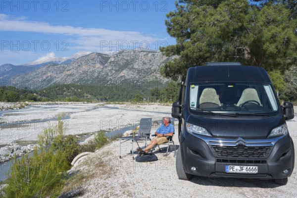 A man sits next to a motorhome on a riverbed surrounded by mountains, camper, Dargaz River near