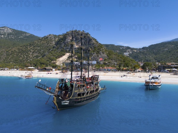 Pirate ship sails in front of a picturesque coastal landscape with mountains and clear blue water,