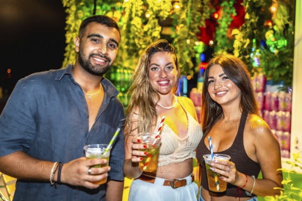 Three friends smiling and holding cocktails at a vibrant outdoor night party, enjoying the festive