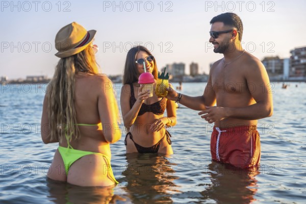Three friends enjoying refreshing cocktails while standing in ocean water at sunset, celebrating