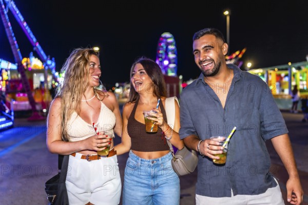 Three friends walking and laughing together at amusement park at night, holding drinks and enjoying